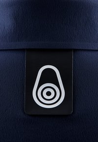 Marineblaue Stoff mit einer strukturierten Oberfläche. Ein rechteckiges schwarzes Etikett zeigt ein weißes Logo, das eine ovale Form und konzentrische Kreise darstellt.