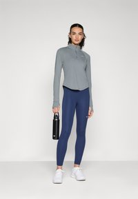 Helles graues Langarmoberteil mit einem Reißverschlusskragen, kombiniert mit marineblauen Leggings. Das Model hält eine schwarze Wasserflasche; weiße Sneakers vervollständigen das Outfit.