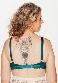 Reggiseno teal con bordatura in pizzo, spalline regolabili e chiusura ad gancio, indossato da una persona con un tatuaggio floreale sulla schiena, pelle chiara e capelli ricci.
