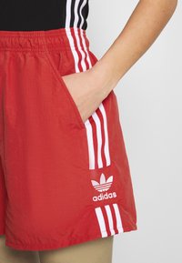 Red athletic shorts with an elastic waistband, white side stripes, and an embroidered white Adidas logo on the lower left side. Pocket visible.