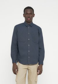 Άντρας ντυμένος με μπλε σκούρο πουκάμισο με κουμπιά και δύο τσέπες στο στήθος, και μπεζ παντελόνι cargo, στέκεται μπροστά σε απλό φωτεινό φόντο.