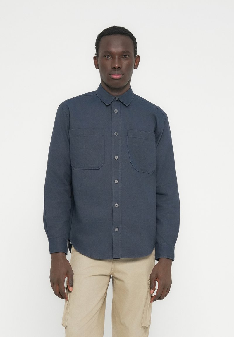 Άντρας ντυμένος με μπλε σκούρο πουκάμισο με κουμπιά και δύο τσέπες στο στήθος, και μπεζ παντελόνι cargo, στέκεται μπροστά σε απλό φωτεινό φόντο.