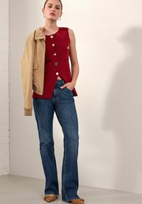 Top rosso senza maniche con bottoni dorati e due tasche frontali, abbinato a jeans svasati blu chiaro e una giacca in suede beige poggiata sulle spalle.