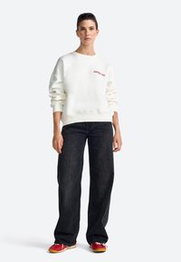 Felpa bianca con "APRES SKI" in rosso, abbinata a jeans neri a gamba larga e scarpe rosse con accenti gialli. Tessuto liscio, design informale.