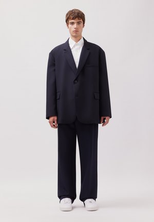 Giovane uomo in piedi che indossa un abito nero oversize, camicia bianca e sneakers bianche su uno sfondo chiaro e semplice.