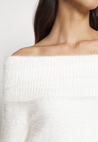 Femme portant un pull blanc duveteux à épaules dénudées, révélant les clavicules et une partie de ses longs cheveux bruns.
