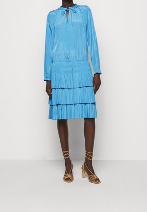 Leuchtend blaues Kleid mit gerafftem Ausschnitt, langen Ärmeln und gestuftem Rock. Glatte Textur mit Schlüsselloch-Ausschnittdetail. Paar beige Riemchen-High Heels.