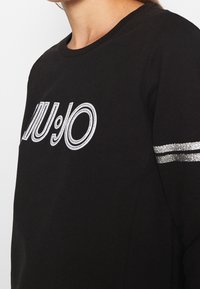 Primo piano di una persona che indossa una maglietta a maniche lunghe nera con lettere contornate in bianco e due strisce di glitter argento sulla manica.