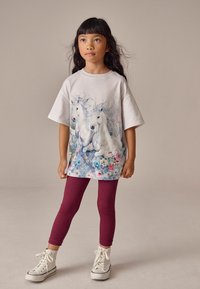 Hellgraues T-Shirt mit einem Einhorn-Graphic und floralem Akzent, kombiniert mit weinrot gerippten Leggings und weißen High-Top-Sneakern.