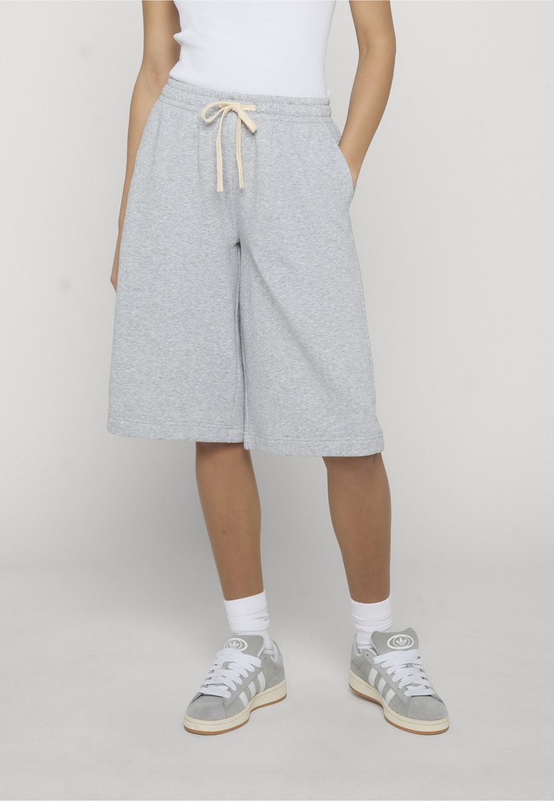 Personne portant un short de sport gris clair mi-cuisses avec cordon de serrage, des chaussettes blanches, des baskets gris et blanc, et un débardeur blanc.
