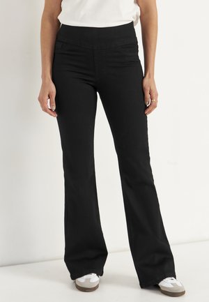 Frau trägt schwarze Bootcut-Hose, weißes Oberteil und weiße Sneakers, steht vor einem schlichten hellen Hintergrund.