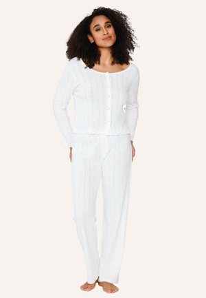 Frau mit lockigem Haar, die eine weiße, lange, knöpfbare Strickjacke und passende Hosen trägt, barfuß vor einem neutralen Hintergrund steht.