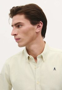 Camicia a righe giallo chiaro con colletto, caratterizzata da un piccolo logo di teschio e ossa incrociate nere sul petto. Vestibilità casual.
