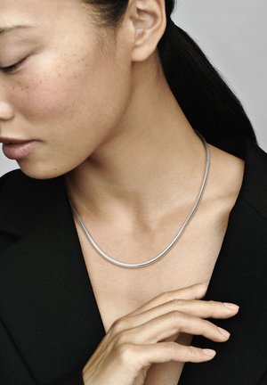Silberne Schlangenkette mit einem glatten, runden Design, die locker um den Hals liegt, kombiniert mit einer schwarzen Kleidung und sichtbarer Hauttextur.