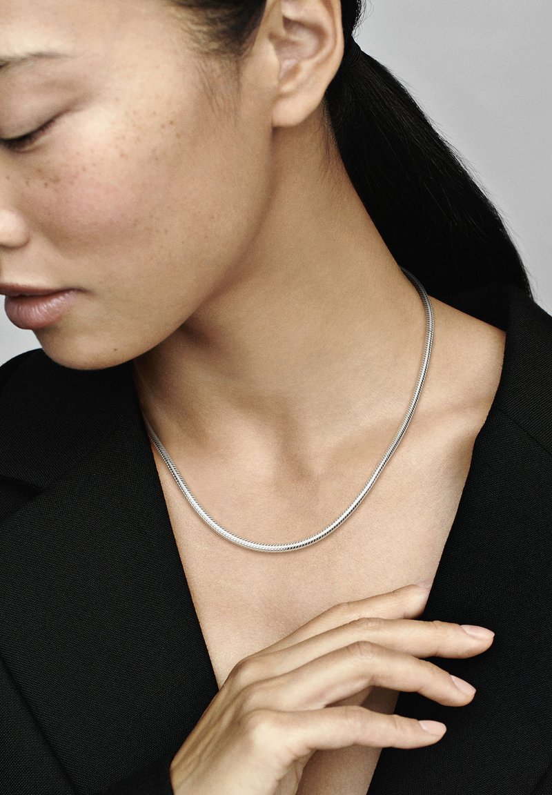 Silberne Schlangenkette mit einem glatten, runden Design, die locker um den Hals liegt, kombiniert mit einer schwarzen Kleidung und sichtbarer Hauttextur.
