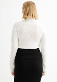 Suéter blanco de punto con un alto cuello y puños acanalados, combinado con una falda negra acanalada, visto desde atrás.