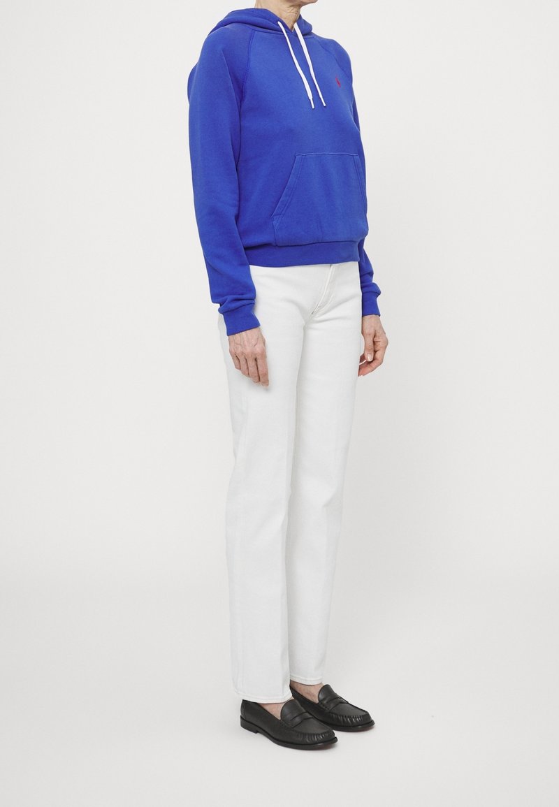 Blauer Baumwoll-Hoodie mit Kängurutasche und weißen Kordeln, kombiniert mit weißen gerade geschnittenen Jeans und schwarzen Slippern.