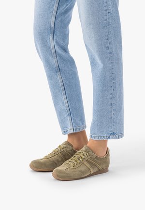 Osoba ubrana w jasnoniebieskie dżinsy i oliwkowo-zielone zamszowe trampki z żółtymi i białymi sznurówkami, stojąca na białym tle.