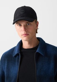 Mlad moški z modrimi očmi, oblečen v črno korduro kapo z napisom "COLMAR," črno srajco in temnobluzom jakno z zadrgo na svetli podlagi.