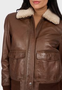 Chaqueta bomber de cuero marrón con cuello de piel sintética, dos bolsillos frontales tipo parche, puños de canalé y una textura suave.