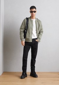 Chaqueta bomber verde oliva, camiseta blanca, jeans negros y botas negras. La chaqueta cuenta con un cierre y bolsillos laterales, con una textura suave.