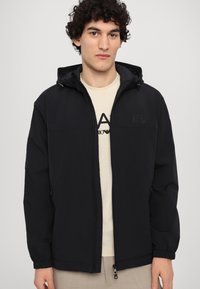 Svart huva jacka med dragkedja fram, elastiska ärmar och en diskret logotypdetalj. Bärs över en ljus färgad t-shirt. Slät tygtextur.