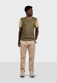 Camisa de manga corta en verde oliva y beige con cuello redondo, que presenta un pequeño logo de elefante. Combinada con pantalones beige y zapatillas grises.