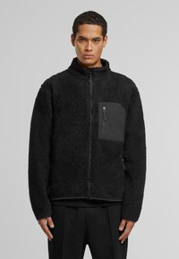 Fekete fleece dzseki magas gallérral, cipzárral záródik, és kontrasztos anyagú mellzsebe van. Strukturált felülete és laza szabása van.