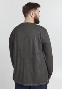 Sötétszürke, hosszú ujjú póló, laza fazonnal, textúrált anyaggal, kerekített aljjal és egy kis márkajelzéssel az oldalon.