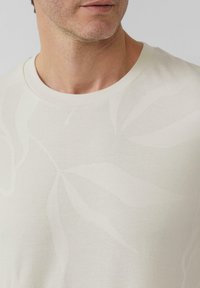 Crèmekleurig shirt met korte mouwen, voorzien van een subtiel loofpatroon en een gestructureerd oppervlak; ronde halslijn en zacht, lichtgewicht materiaal.