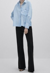 Camicia over-size a righe blu chiaro con taschino sul petto, abbinata a pantaloni svasati neri e scarpe aperte, su uno sfondo neutro.