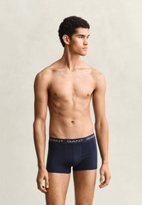 Jeune homme debout pieds nus, torse nu, portant un boxer bleu marine foncé avec le logo "GANT" sur la ceinture, regardant de côté sur un fond uni.