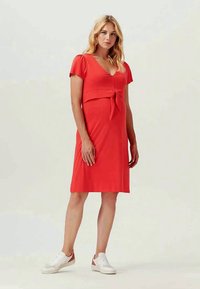 Vestido casual rojo con un corsé ajustado, mangas cortas abullonadas y un detalle de lazo en el frontal. Textura suave, hasta la rodilla, combinado con zapatillas blancas.