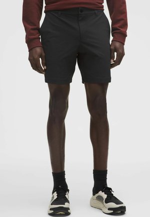 Sorte shorts med fladt frontstykke og bæltestropper, kombineret med sorte og cremefarvede atletik-sko. Stoffet ser glat ud med en skræddersyet pasform.