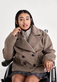 Jeune femme en fauteuil roulant portant un manteau marron et une jupe à carreaux, reposant une main sur l'accoudoir du fauteuil roulant et l'autre près de son visage.