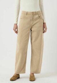 Osoba v béžových širokých kalhotách, bílém dlouhém rukávu a hnědých semišových botách na klínku stojící na jednobarevném pozadí.