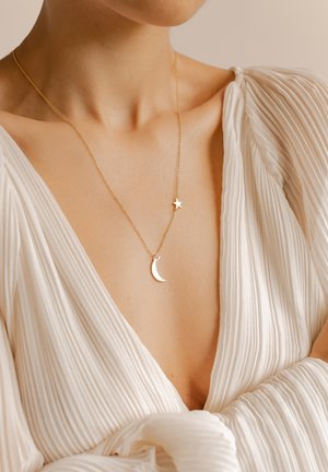 Gros plan d'une personne portant un chemisier blanc plissé et un collier en or avec des pendentifs en forme de croissant de lune et d'étoile sur la clavicule.
