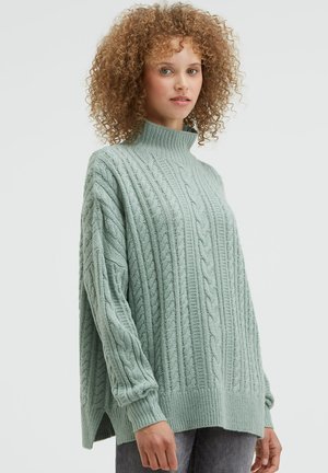 Light green cable-knit sweater with a high collar, long sleeves, ribbed cuffs, and side slits. Made of soft, textured material.