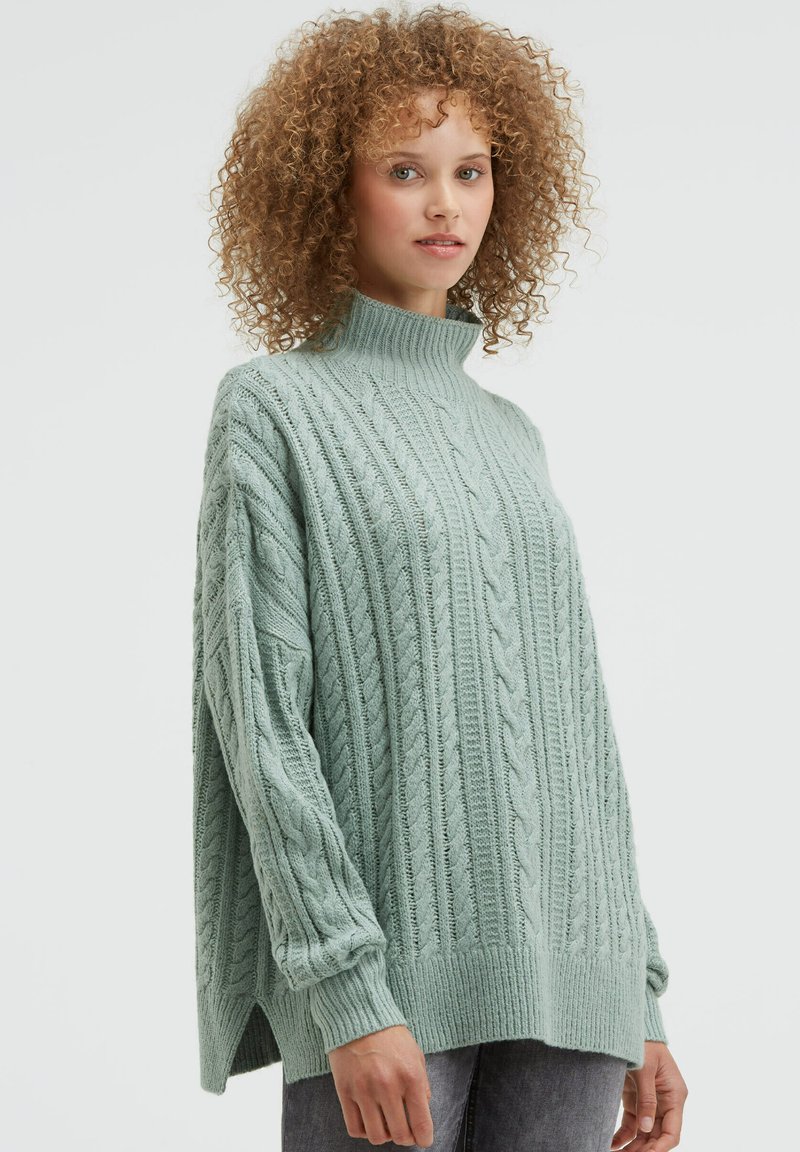 Hellgrüner Strickpullover mit hohem Kragen, langen Ärmeln, gerippten Bündchen und seitlichen Schlitzen. Hergestellt aus weichem, strukturiertem Material.