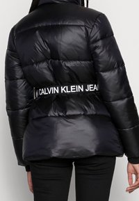 Černá pufová bunda s lesklým povrchem, s vodorovným šitím a výrazným bílým logem v pase.