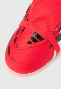 Κοντινό πλάνο δαχτύλου κόκκινου αθλητικού παπουτσιού με μαύρες λεπτομέ ρειες και υφασμένο λουράκι με μαύρο λογότυπο Adidas.