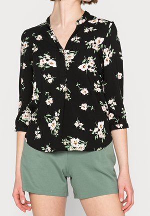 Kvinde iført en sort blomstret bluse med lange ærmer og grønne højtaljede shorts, stående foran en ensfarvet baggrund.