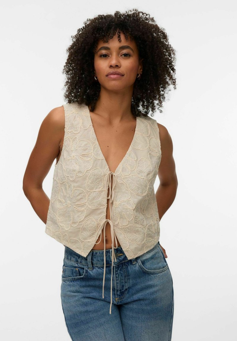 Femme aux cheveux bouclés portant un gilet beige sans manches avec broderie florale et un jean bleu, mains dans le dos sur fond blanc.