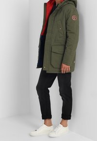 Persona vestida con un abrigo con capucha color verde oliva y forro rojo, pantalones negros enrollados y zapatillas blancas, de pie contra un fondo liso.
