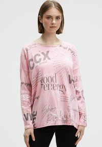 Junge Frau mit kurzem braunem Haar, die ein lockeres rosa Langarmshirt mit verschiedenen motivierenden Sprüchen und schwarze Hosen trägt.