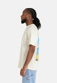 Korte mouwen, crèmekleurig t-shirt met een kleurraf beach scene graphic op de achterkant. Gemaakt van zachte katoen met een relaxte pasvorm.