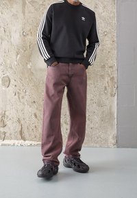 Man i svart Adidas sweatshirts, blekta mauve raka jeans och svarta skulpturerade sneakers står mot en texturerad vägg.