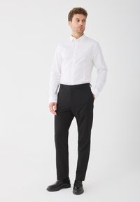 Férfi fehér gombos ingben, fekete nadrágban és fekete cipőben áll, egyik kezét a zsebében tartva, fehér háttér előtt.