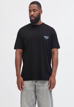 Man with beard wearing black Bushwick t-shirt and light gray jeans stands against plain white background, looking down to his left.