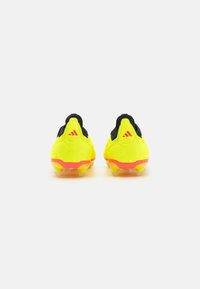 Botas de fútbol amarillas con un acento negro, que presentan un material texturizado, un diseño de tacón acampanado y taquetes translúcidos. Marca roja en el talón.